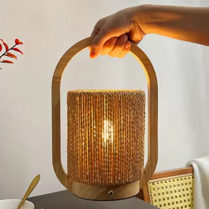 A hand holds the handle of a table lamp with a natural woven rattan shade and a light wood frame, lit with a warm glow against a softly blurred indoor background.