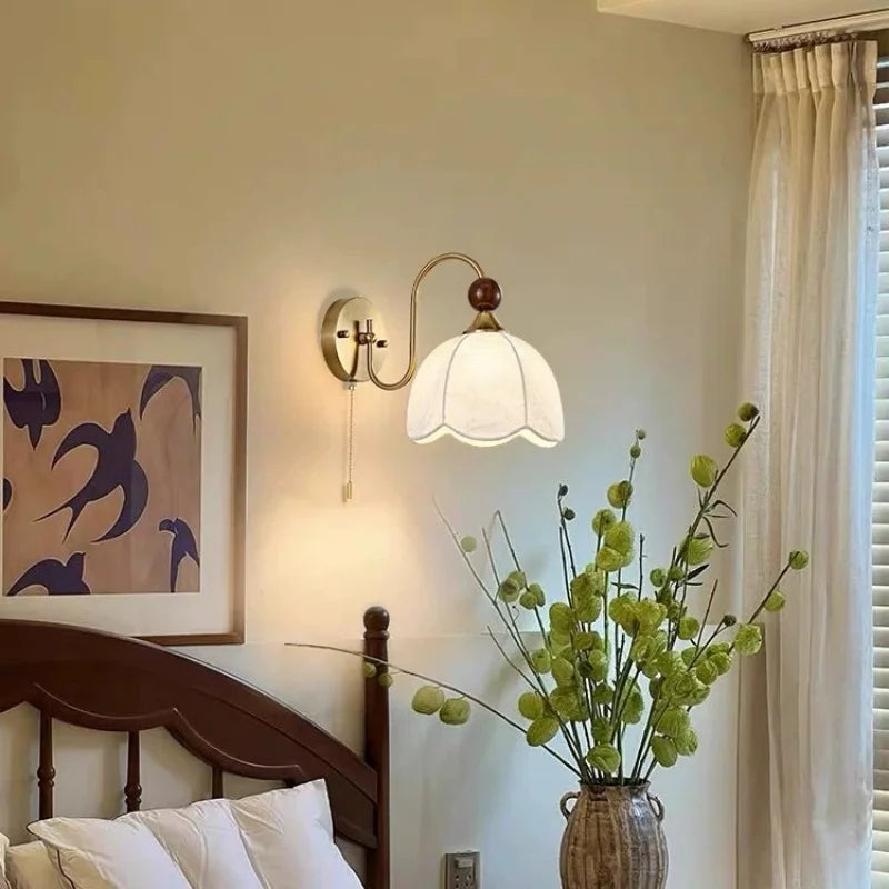 A wall-mounted sconce with a white bell-shaped glass shade and brass-toned metal arms, installed above a bed in a softly lit bedroom. A framed abstract print, wooden headboard, white bedding, and a vase with green stems are visible.