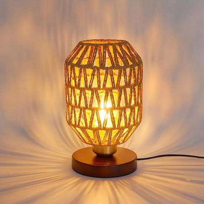 A warm-toned woven table lamp with a geometric patterned shade and a round wooden base, glowing with a soft yellow light against a softly lit neutral background.