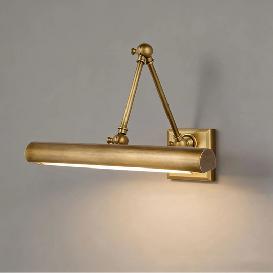 A wall-mounted cylindrical light fixture with adjustable articulated arms, finished in a warm brushed brass or gold tone, shown lit against a plain gray wall.