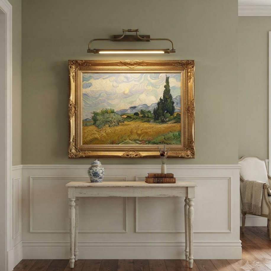 A framed landscape painting hangs on a sage green wall above a white console table in a styled room. The painting depicts a golden field, green trees, and a cloudy sky, enclosed in an ornate gold frame.