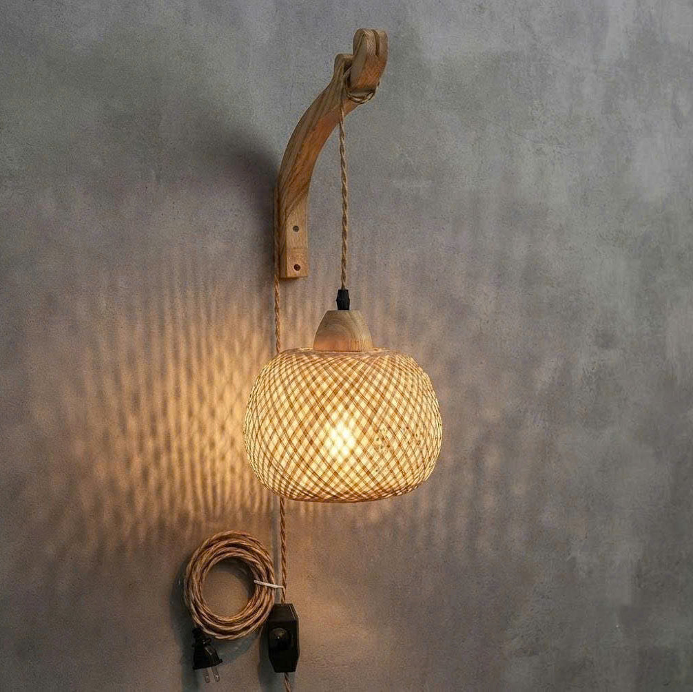 A rustic wall-mounted pendant light with a curved natural wood backplate, a woven rattan shade, and a hanging cord with a plug, shown against a textured gray wall.