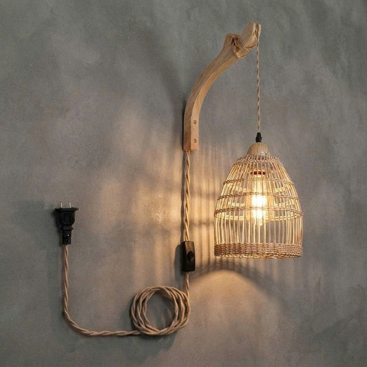 A rustic wall-mounted pendant light with a curved natural wood branch arm, a woven rattan birdcage shade, and a coiled beige cord with a black plug, shown against a textured gray wall.