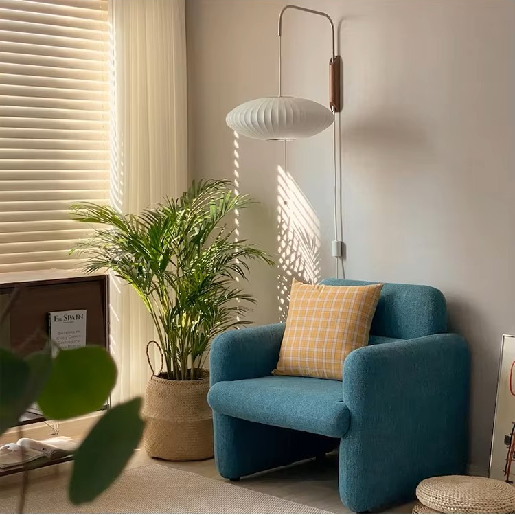A cozy interior corner featuring a teal blue armchair with a yellow-and-white checkered accent pillow, next to a potted green plant and a white floor lamp in warm natural light.