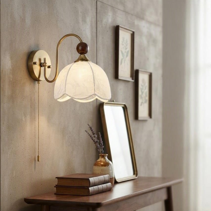 A vintage-style wall sconce with a warm brass finish and a white fabric flower-shaped shade, mounted on a textured beige wall above a wooden console table with books and a mirror.