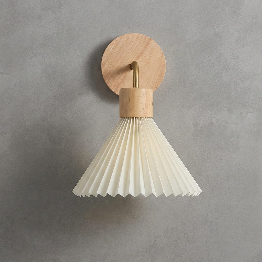 A modern wall-mounted light fixture with a circular natural wood backplate, a brass-toned central rod, a cylindrical wood cap, and a pleated white paper or fabric shade, shown against a textured gray wall.