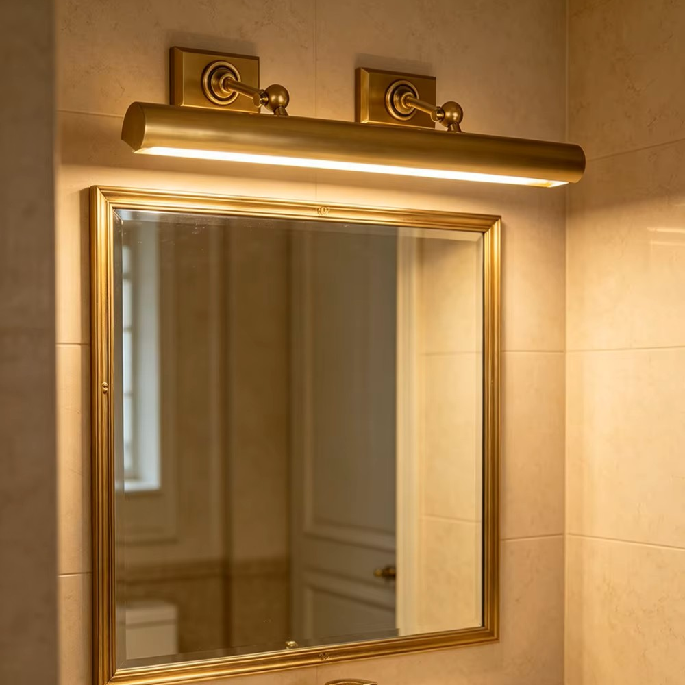 A gold-framed bathroom mirror mounted on a beige tiled wall, illuminated by a matching gold wall sconce with a cylindrical light bar.