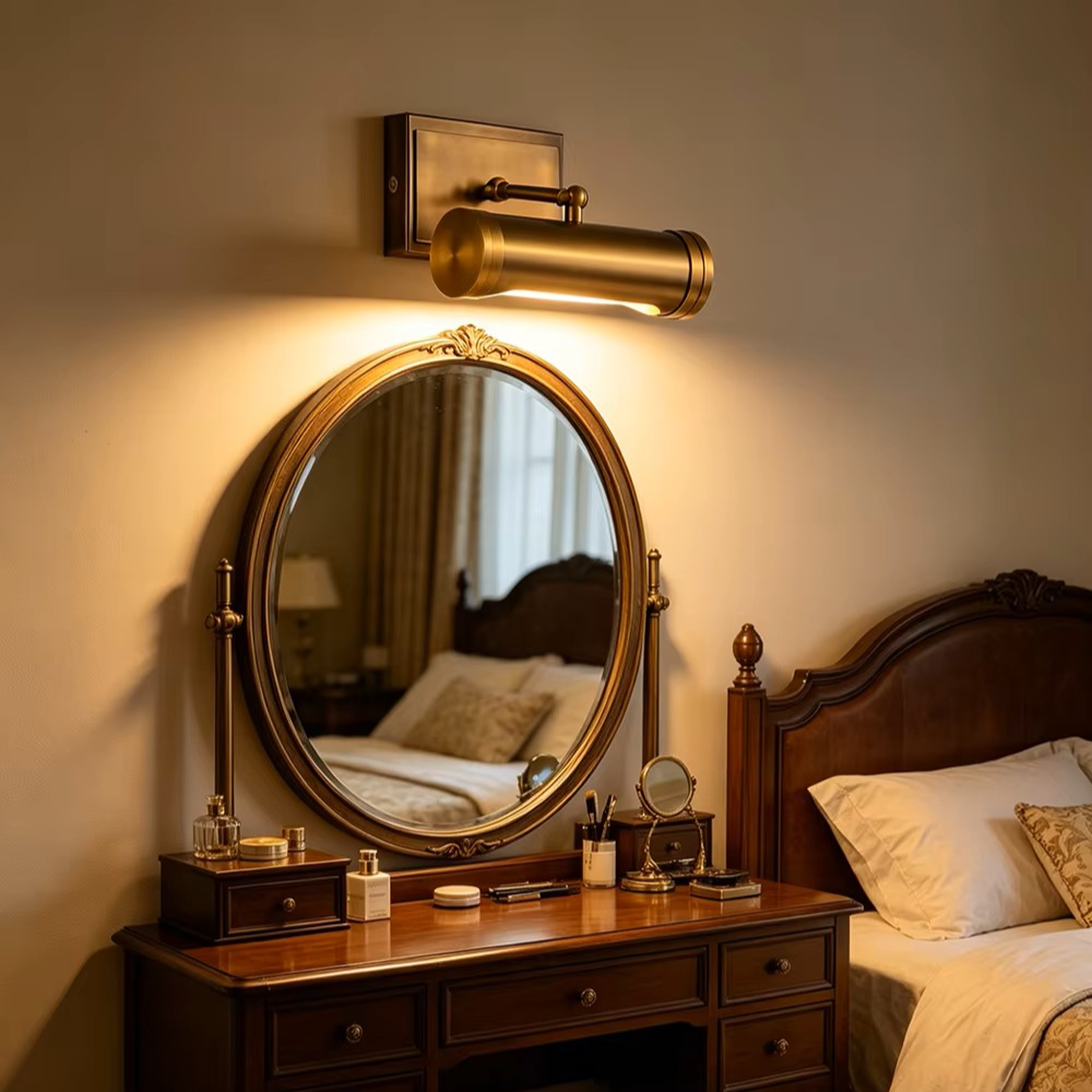 A brass wall-mounted cylindrical sconce with a warm glowing light, mounted above an ornate round vanity mirror on a bedroom wall.