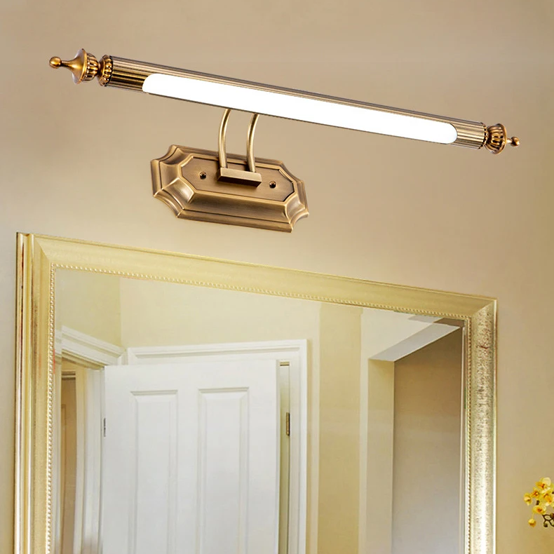 An antique bronze bathroom light fixture with a curved bar and decorative wall mount, shown installed above a gold-framed mirror in a beige bathroom setting.
