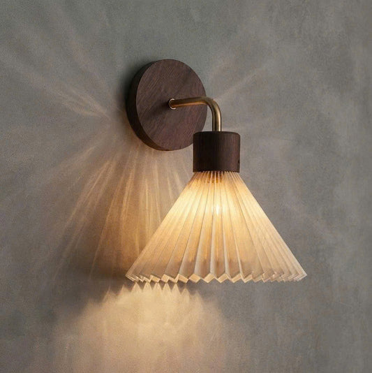 A modern wall sconce light fixture with a pleated clear glass shade, a dark brown wood accent, and a brass-colored curved arm, mounted on a textured gray wall and casting a warm light.