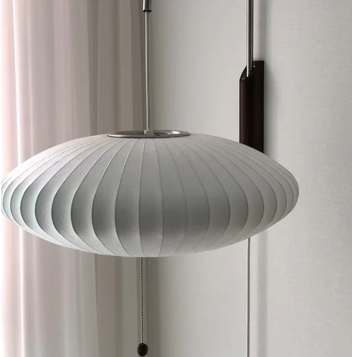 A modern pendant light with a ribbed, pleated white shade suspended from a slim metal rod, shown against a neutral off-white wall and curtain background.