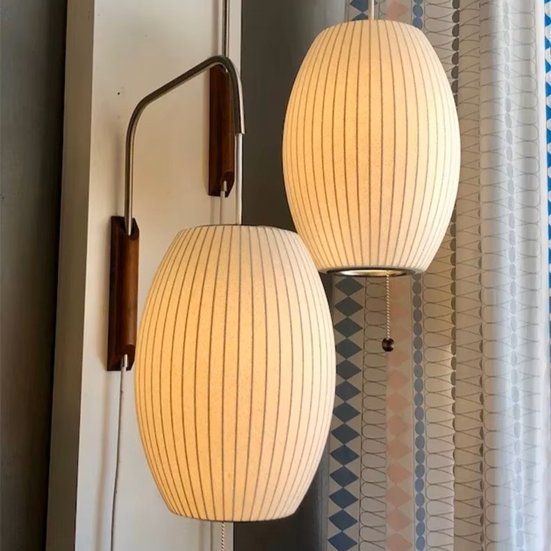 Two modern wall-mounted pendant lights with ribbed cream fabric shades and curved dark metal arms, shown hanging in front of a patterned curtain.