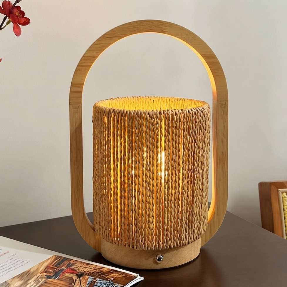 A lit table lamp with a natural bamboo frame and a cylindrical woven rattan shade, placed on a dark tabletop against a light wall.
