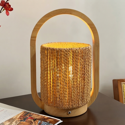A lit table lamp with a natural bamboo frame and a cylindrical woven rattan shade, placed on a dark tabletop against a light wall.