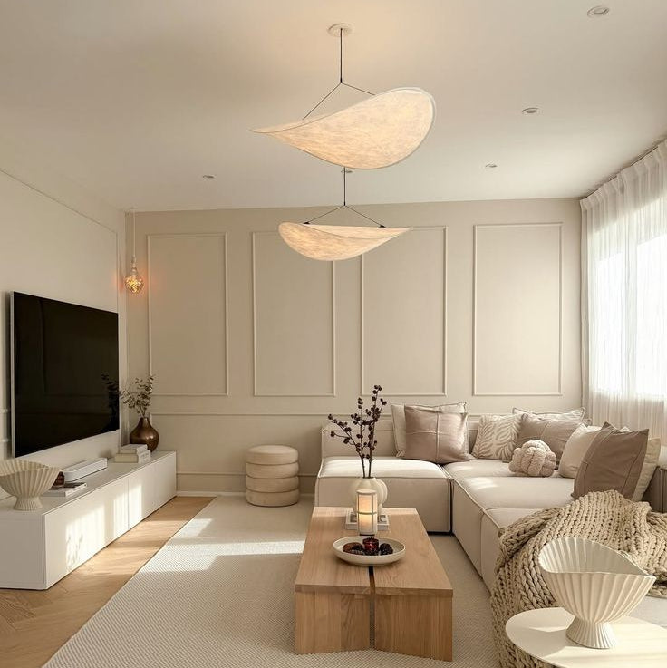 A modern, minimalist living room featuring a light beige sectional sofa, a low wooden coffee table, a large textured cream rug, and a white media console with a television. The room is styled with warm neutral tones, soft natural light, and contemporary decor. wabi_sabi_pendant_light_wabi_sabi_chandelier_living_room_pendant_light_wabisabi_lighting_ideas_wabi_sabi_living_room