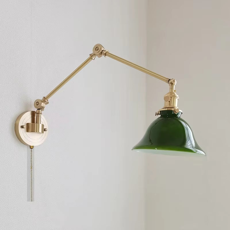 A wall-mounted adjustable desk lamp with a brass-colored metal arm and circular wall plate, featuring a glossy dark green glass shade and a pull chain.