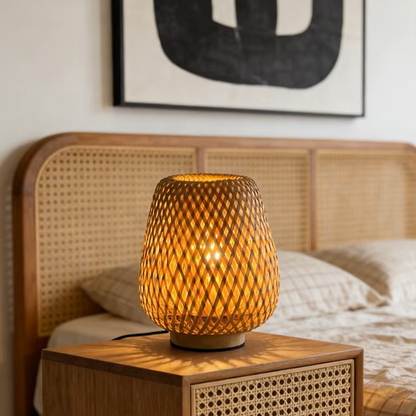 A warm glowing woven rattan table lamp sits on a wooden bedside table in a bedroom setting with a cane headboard and neutral bedding.