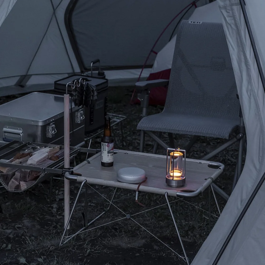 A gray folding camp table set up inside a tent at night, with a lit lantern, bottle, and small round container on top, alongside a gray folding chair and camping gear.