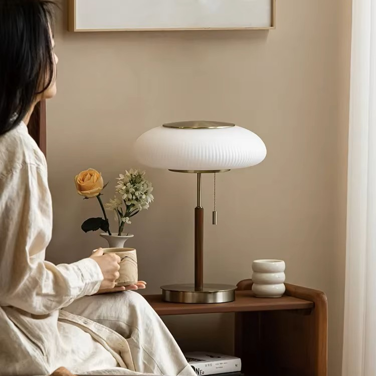A modern table lamp with a ribbed white oval shade, a dark brown wooden stem, and a brushed brass base sits on a wooden bedside table in a cozy room. A woman in a beige robe is seated on the left, holding a mug, with a small vase of flowers and stacked ceramic cups on the table.