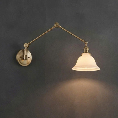 An adjustable wall-mounted swing arm sconce with an antique brass finish and a glowing white glass bell shade, mounted on a dark gray wall.