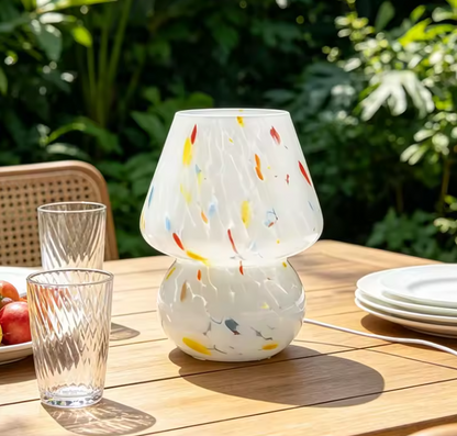 A modern two-tier table lamp with a white frosted glass shade and base, decorated with colorful speckles of red, yellow, and blue, set on a wooden outdoor dining table with glassware and plates.