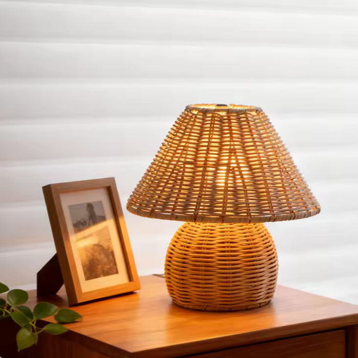 A warm-toned woven rattan table lamp with a conical shade and rounded base, placed on a wooden side table next to a framed picture against a white slatted background.
