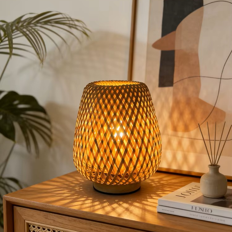 A warm glowing table lamp with a woven bamboo or rattan cylindrical shade sits on a wooden side table in a styled room. A framed abstract print, a small vase with reeds, and a book are nearby, with a plant leaf visible on the left.