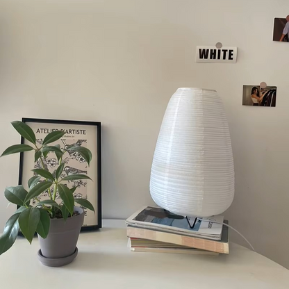 A ribbed white table lamp shaped like a teardrop sits on a stack of books on a white table, next to a potted green plant and a framed print, against a beige wall with small decorative pins.