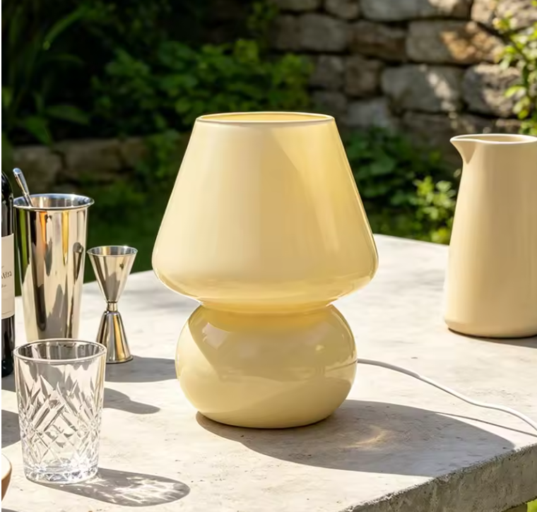 A pale yellow table lamp with a rounded, bulbous design sits on a stone table outdoors, surrounded by bar and serving props like a cocktail shaker, glass, and pitcher.
