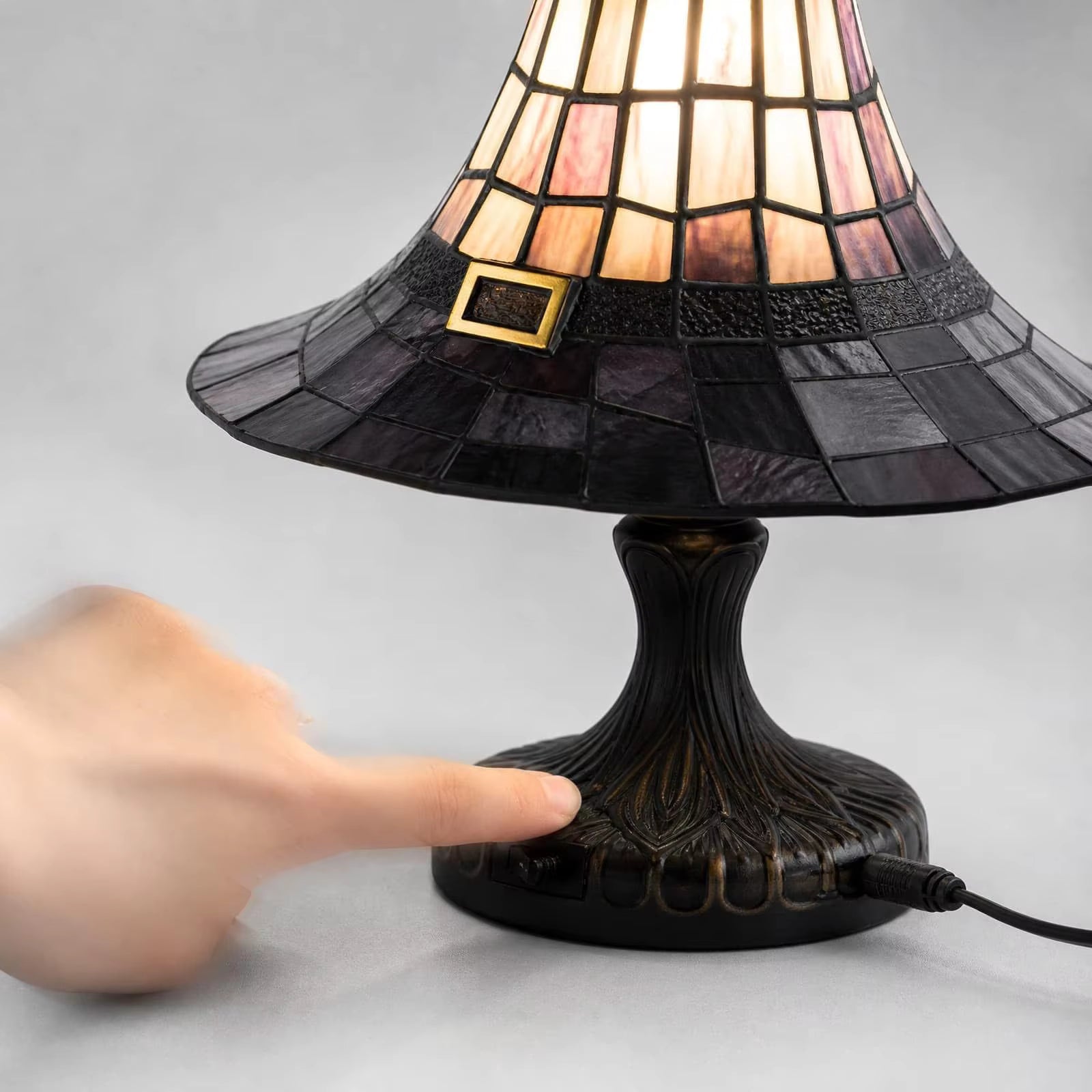 A stained-glass table lamp with a black sculpted base and a conical shade made of pink, purple, and black glass panels, shown with a hand pressing the base to turn it on.