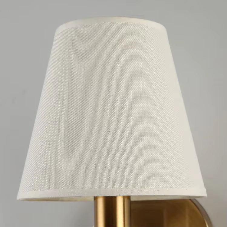 A close-up of a table lamp shade made of textured off-white fabric, mounted on a brushed gold metal base against a neutral gray wall.