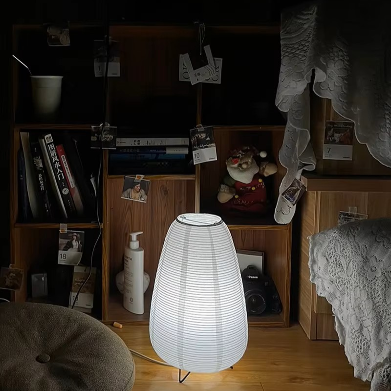 A glowing white paper lantern table lamp with a ribbed conical shade stands on a wooden floor in a dimly lit bedroom or study setting with shelves, books, and bedding visible in the background.