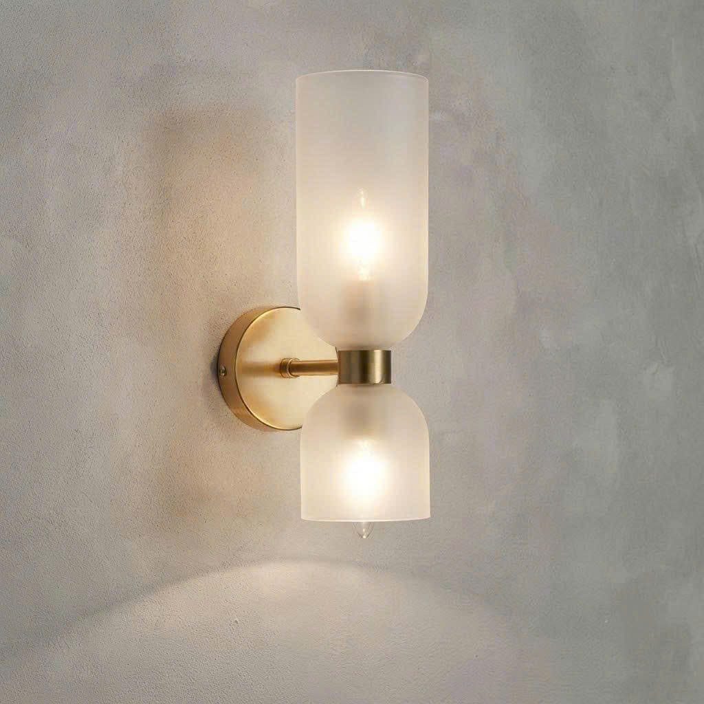 A modern double-shade wall sconce with frosted glass shades and a brushed brass metal mount, shown illuminated against a textured gray wall.