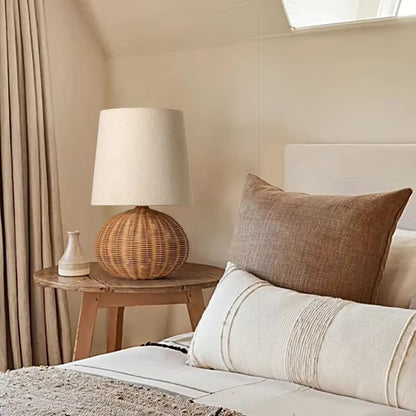 A woven rattan table lamp with a cream fabric drum shade sits on a round wooden nightstand beside a bed with neutral beige and cream pillows in a softly lit bedroom.