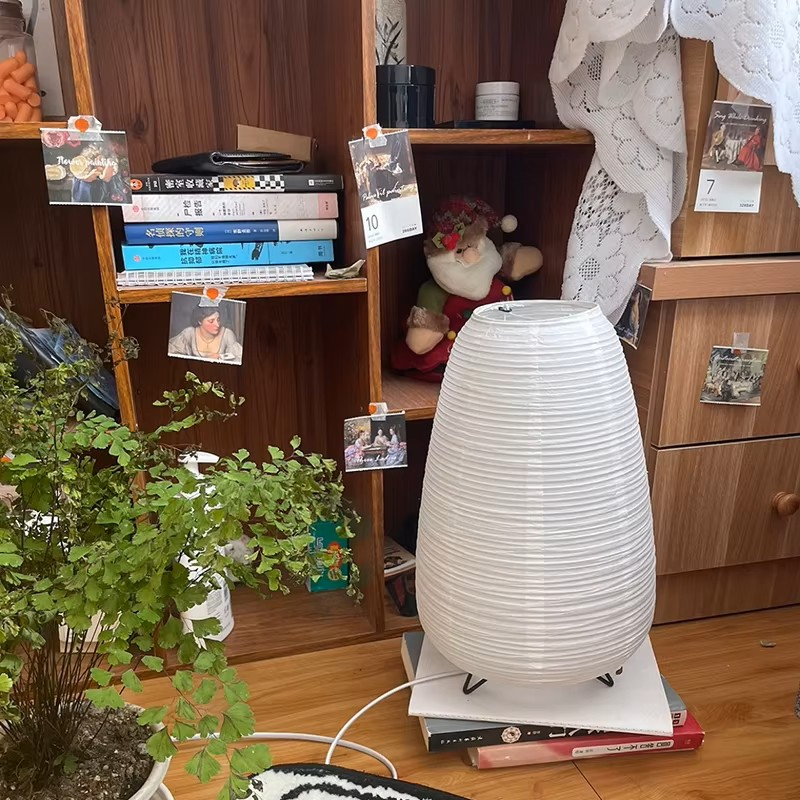 A white ribbed paper lantern-style table lamp sits on a stack of books on a wooden floor in front of a wooden shelf unit filled with books, photos, and a plush toy.
