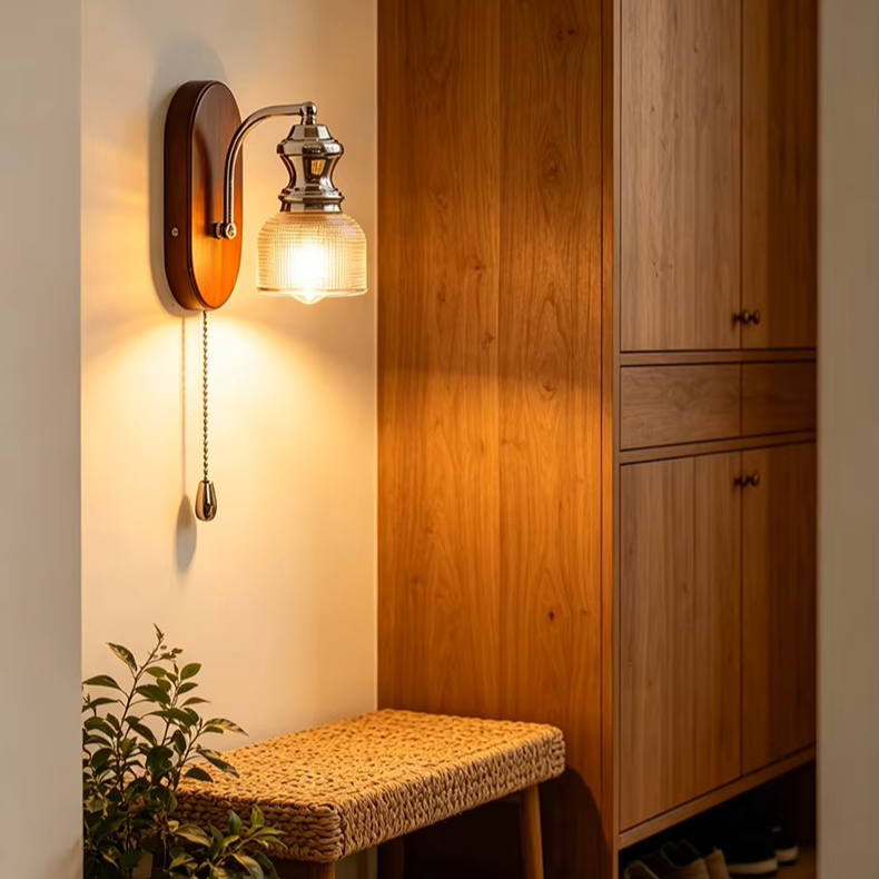 A warm wall-mounted sconce with a pull chain, shown lit in a real interior setting beside a wooden cabinet and bench.