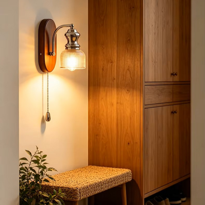 A warm wall-mounted sconce with a pull chain, shown lit in a real interior setting beside a wooden cabinet and bench.