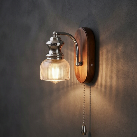 A vintage-style pull-chain wall sconce with a ribbed clear glass shade, polished silver metal hardware, and a curved wooden backplate, mounted on a textured dark gray wall and casting a warm glow.