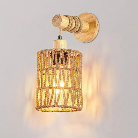 A rustic wall-mounted light fixture with a cylindrical woven rope shade, natural wood wall plate and arm, and a glowing warm bulb inside, shown against a light gray wall.
