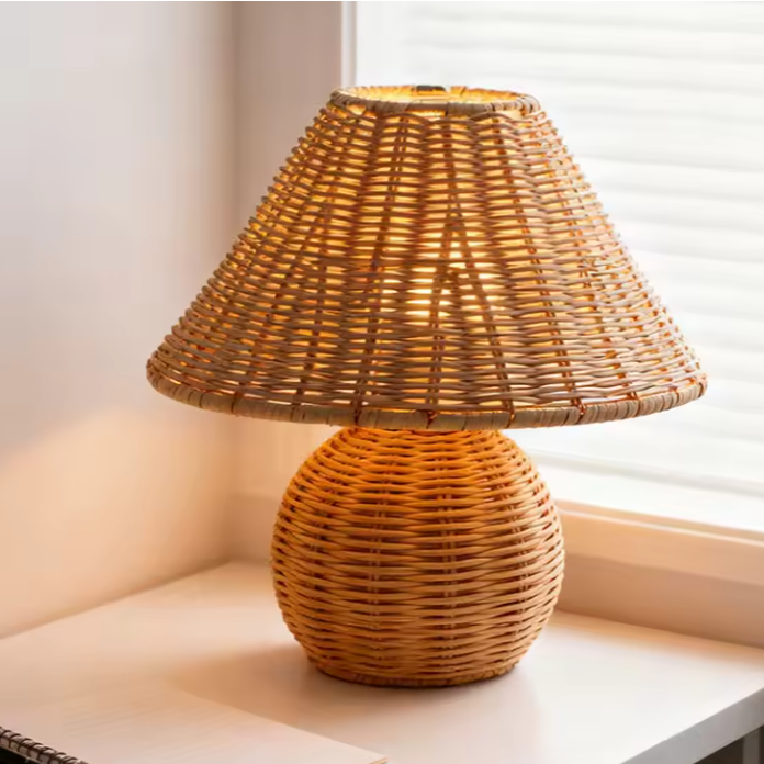 A natural woven rattan table lamp with a conical shade and a round textured base, shown on a white windowsill with soft window light.