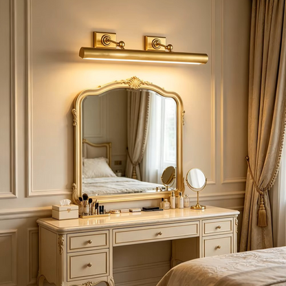 A classic gold-framed vanity mirror with an integrated horizontal wall light above it, set in a luxurious cream-colored bedroom with beige curtains and a bed visible in the reflection.