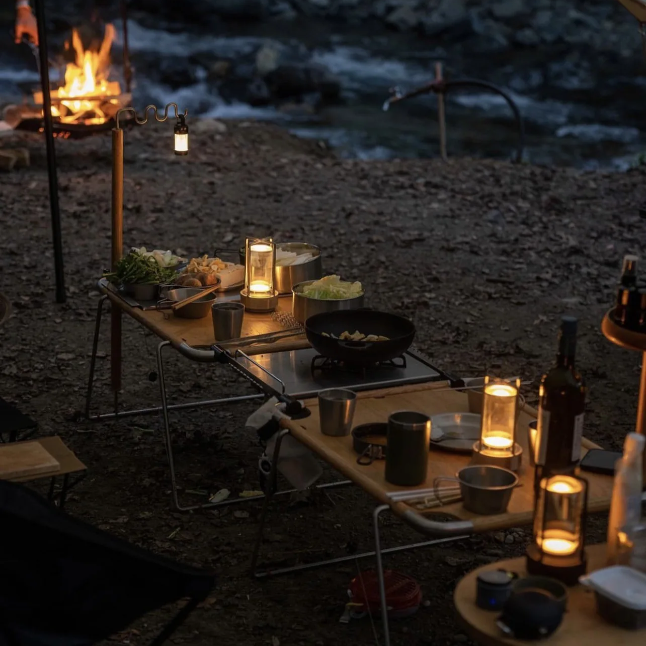 Outdoor camping cooking setup at dusk by a river, with folding tables, lanterns, a portable stove, bowls, and a campfire in the background.
