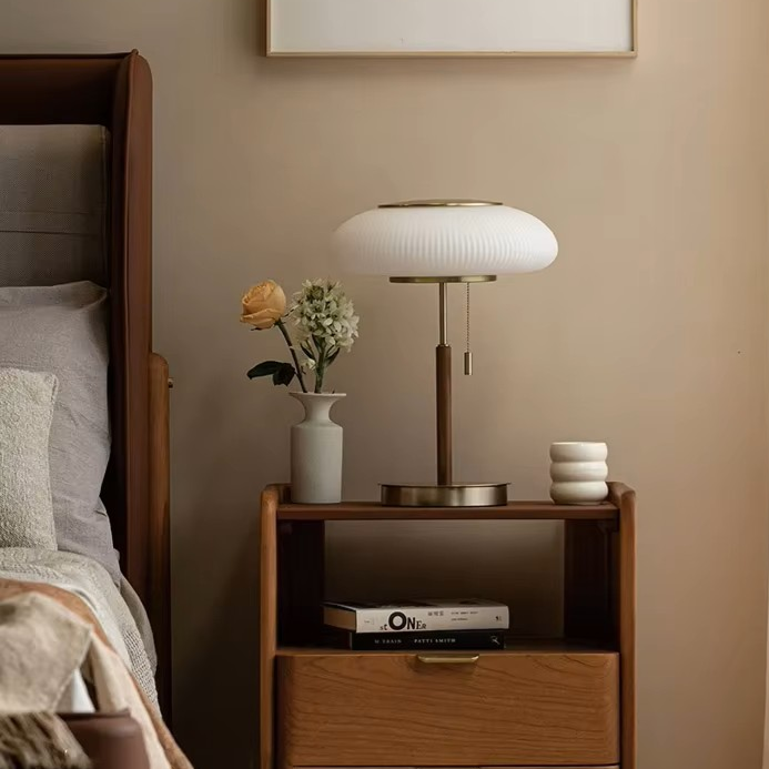 A modern table lamp with a ribbed white oval shade and brushed brass base, styled on a wooden nightstand in a warm beige bedroom setting.