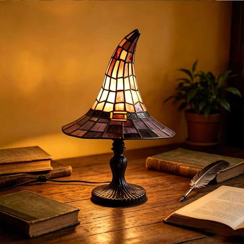 A stained-glass-style table lamp shaped like a witch hat sits on a wooden desk, glowing warmly. The lamp has a dark metal base and a conical shade made of amber, orange, and brown glass panels.