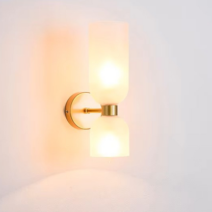 A modern double-light wall sconce with frosted cylindrical glass shades and a brushed brass metal mount, shown illuminated against a plain white wall.