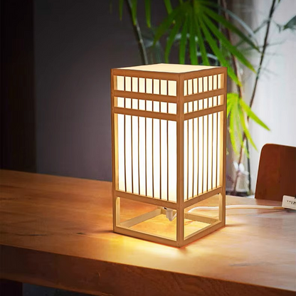 A Japanese-style wooden table lamp with a warm glowing light, featuring a square frame and vertical slatted panels, sitting on a wooden table in a room setting.