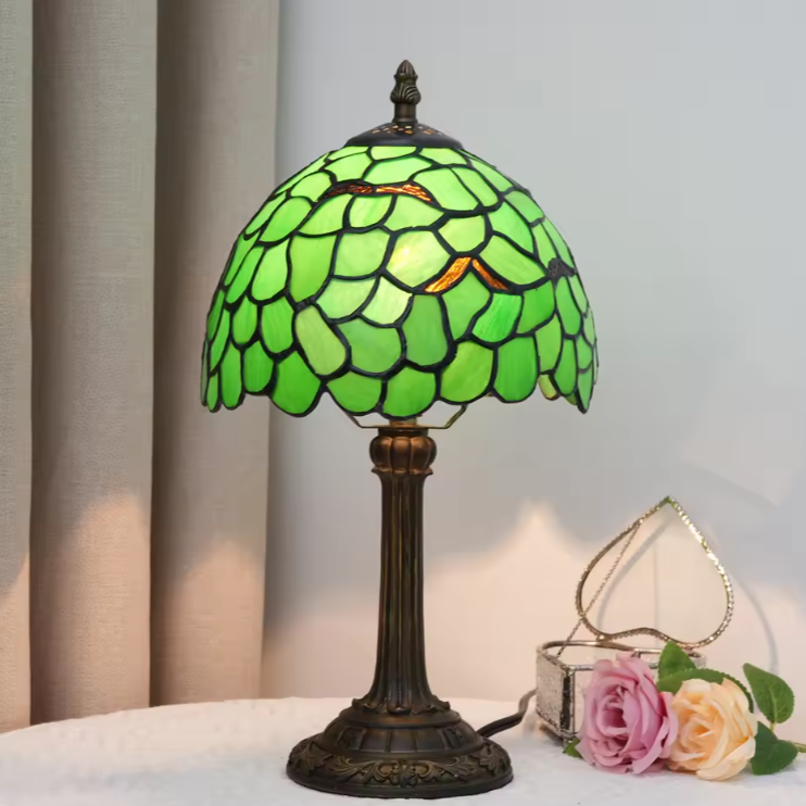 A Tiffany-style table lamp with a green stained-glass shade and a dark bronze metal base, shown on a white surface next to pink and cream roses against a light gray curtain background.
