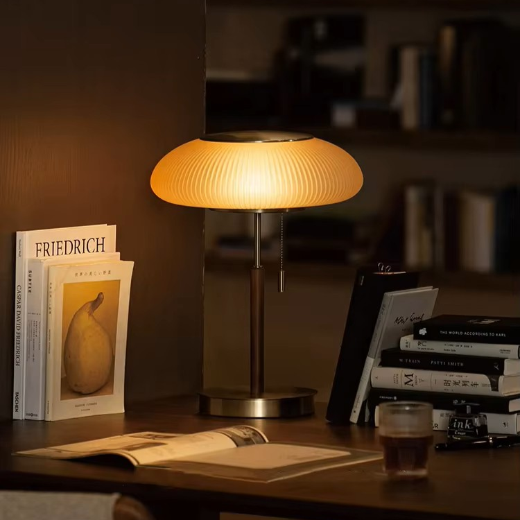 A modern table lamp with a ribbed, mushroom-shaped shade glowing with warm light, set on a dark wooden desk next to books and an open notebook in a cozy room setting.