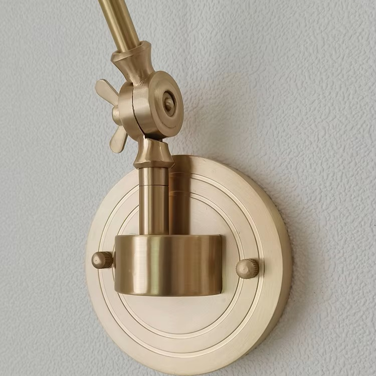 A close-up of a brass wall-mounted light fixture with an adjustable arm and circular backplate, shown against a textured off-white wall.