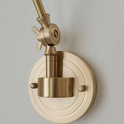 A close-up of a brass wall-mounted light fixture with an adjustable arm and circular backplate, shown against a textured off-white wall.
